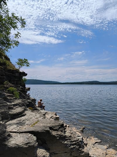 The image portrays an outdoor natural landscape in Horaivka, Ukraine. The scene features a vast body of water, likely a lake or wide river, extending across the midground and background, displaying light surface ripples. On the left side and in the foreground, a multi-layered, grey and brown rocky cliff face descends towards the water, with green vegetation, including small trees and bushes, growing on its ledges and summit. The distant horizon across the water is marked by dark green, forested hills. The sky above is clear blue with scattered white cirrocumulus and altocumulus clouds. A single adult male, with light-colored hair and wearing patterned shorts, is seated on a flat rock ledge at the cliff's base, close to the water's edge, positioned with his back partially towards the viewer. His head is tilted downwards, indicating engagement with an unseen object held in his hands or focused on the water immediately in front of him. No other individuals or discernible objects are present in the immediate vicinity of the male.
