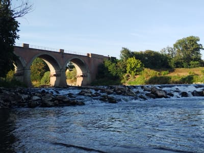 A red brick viaduct with arched openings spans across a river. The river flows over rocks, creating a small waterfall. Lush green trees and vegetation line the riverbanks and the area behind the viaduct. The sky is clear and blue. The scene appears to be located in the city of San Colombano al Lambro, Italy, near the Lambro river.
