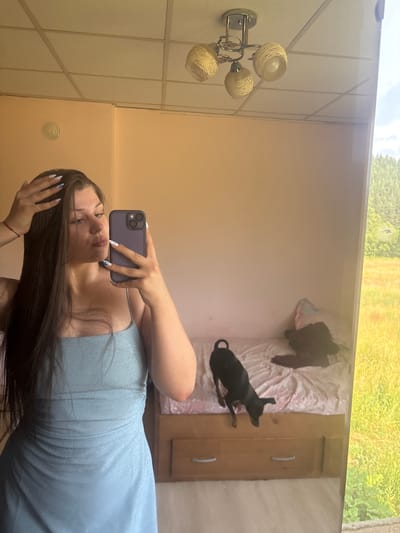 This media file is a mirror selfie taken by a young woman. She is wearing a light blue, sparkly dress and has long, dark hair. Her nails are long and decorated with white and blue designs. She is holding a dark purple smartphone to take the picture.
The reflection in the mirror shows a bedroom. There is a bed with white and pink bedding, and a small black dog is lying on the bed. Clothes, including a dark top, are scattered on the bed. To the right of the bed, there is a wooden dresser with two drawers. The walls of the room are painted a light peachy color. On the left side of the reflection, a wall-mounted light fixture with three globe-shaped lamps is visible. Above the wall, a reflection of the sky and trees can be seen, suggesting it is daytime and likely outdoors or near a window.
The scene appears to be a casual moment captured indoors, possibly in a home in Prodanovtsi, Bulgaria, given the location context. The woman's pose and expression suggest a playful or self-aware mood. There is no visible text in the image.