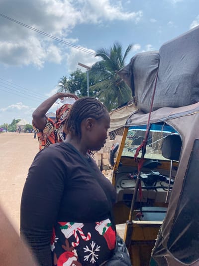 An outdoor scene in Nguzu Edda, Nigeria, depicts a woman from the chest up, facing right and looking down. She has dark skin and braided hair, wearing a dark long-sleeved shirt and a black skirt with a Christmas-themed pattern of snowflakes, reindeer, and candy canes. To her left, another person, also with dark skin and braided hair, is visible from the shoulders up. This person is wearing an orange, yellow, and black patterned garment and has their right arm raised, holding a clear plastic container on their head. They are looking to their left. In the background, a yellow and tan-colored auto-rickshaw, also known as a "keke," is parked. The vehicle is partially covered with a large, grey, textured tarp. A red ribbon or cord hangs from a pole near the auto-rickshaw's windshield. The ground is a dusty, light brown surface, and the sky above is blue with scattered white clouds. Palm trees and other vegetation are visible in the distance, along with some buildings and other people walking or on bicycles.