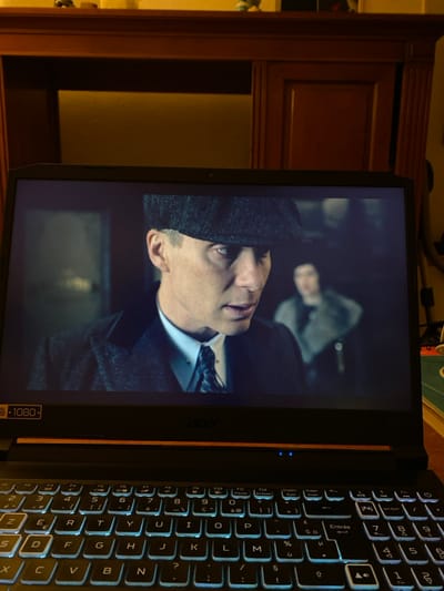 This image captures a black Acer laptop displaying a scene from a television series or film, positioned indoors in Bonnieux, France. The laptop features a backlit QWERTY (French AZERTY) keyboard with light blue illumination and a screen bezel indicating "1080p" resolution. The screen shows a male character, identified as Cillian Murphy's character Thomas Shelby from "Peaky Blinders," prominently in the foreground. He wears a dark tweed flat cap, a dark overcoat, a white collared shirt, and a patterned tie with a tie pin. His expression is intense, with a direct, serious gaze. A second, blurry individual, possibly female and wearing a light-colored, fur-collared garment, is visible in the darker background to the right of the primary character. Behind the laptop, a substantial piece of dark brown wooden furniture, possibly a cabinet or hutch, occupies the upper portion of the frame. It features carved panels and is dimly lit, with a warm, yellowish glow emanating from behind recessed sections. Small, indistinct objects are present on the top surface of the furniture. The overall environment suggests an indoor setting with low ambient lighting, focusing on the screen's content.