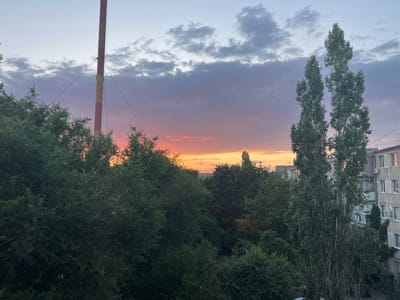 This is a photograph of a cityscape in Azov, Russia, taken during sunset. The sky is filled with a gradient of colors, from pale blue at the top to vibrant pink and orange near the horizon, with scattered grey clouds. Prominently featured in the left portion of the image is a tall, red and white striped smokestack with a ladder running up its side, supported by guy wires. In the foreground and middle ground, a dense canopy of various green trees obscures much of the ground. Two tall, slender trees with sparse foliage stand in the right-middle of the frame. To the right, the edge of a multi-story brick apartment building is visible, with windows, balconies, and air conditioning units. The overall scene depicts an urban environment juxtaposed with natural elements under a colorful evening sky. There are no people or identifiable actions depicted in the image.