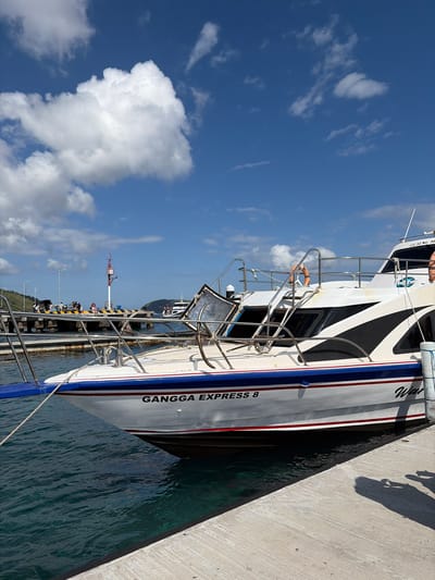 The image shows a white boat with black and blue accents docked at a pier in Kampung Toyapakeh, Indonesia. The boat is named "GANGGA EXPRESS 8" in black lettering. The pier is made of concrete and is lined with bollards. There are several people on the pier, some of whom appear to be waiting to board or disembark the boat. The water is a clear turquoise color. In the background, there are lush green hills and a bright blue sky with fluffy white clouds. The scene is peaceful and serene, with a sense of calm and tranquility. The overall impression is one of a beautiful day at a tropical paradise.