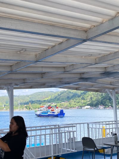 A woman with dark hair pulled back is standing on a deck, looking to the left with her arms crossed. She is wearing a black t-shirt. Behind her, a ferry with a blue hull and white and red upper decks is moving across a body of water. The water is calm and reflects the blue sky. In the background, a lush green hillside dotted with houses and trees slopes down to the water. The scene appears to be in the city of Parapat, Indonesia, likely on Lake Toba. The foreground of the image is dominated by the overhead structure of the ferry deck, with its metal beams and corrugated ceiling. A black plastic chair and a table are also visible on the deck.