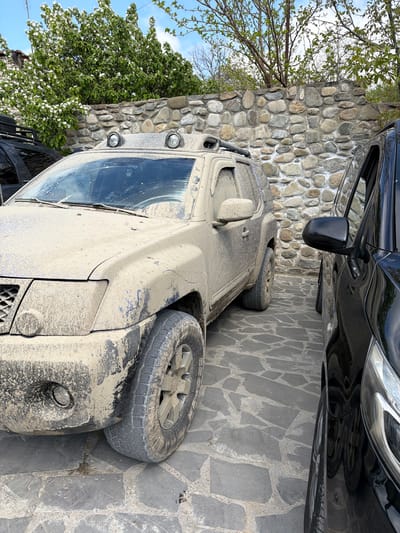A close-up shot depicts a heavily mud-covered, light-colored SUV parked alongside a black vehicle and a cobblestone wall. The SUV is positioned diagonally in the frame, with mud splattered across its front, side, and tires. A portion of a black vehicle, including its side mirror, is visible above the SUV. To the left, a wall constructed of rounded stones is partly obscured by foliage. The ground is covered with irregular cobblestone pavers. The location is Manavi, Georgia.