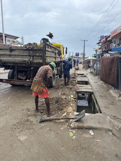 The image captures an outdoor scene in Lagos, Nigeria, likely during daylight hours with overcast skies. Two men are engaged in work near a yellow truck that is partially loaded with soil. One man, wearing a green cap and a blue t-shirt with the number "03" on the back, stands near the front of the truck. The other man, also wearing a green cap and an orange and blue patterned shirt with coral shorts, is positioned at the rear of the truck, appearing to be loading or unloading the soil.
The setting is a dirt and concrete area, with a drainage channel running alongside the path. Debris and litter are scattered across the ground, suggesting a busy urban environment. In the background, a row of buildings and shops line the street, with some signage visible. Power lines and utility poles are also present, typical of an urban landscape. A shovel lies on the ground near the drainage channel. The overall atmosphere suggests manual labor in a working-class area. There is no clear indication of emotion, but the activity points to a task in progress. Some visible text includes "SMOOSKY TRADERS" on a building in the background.