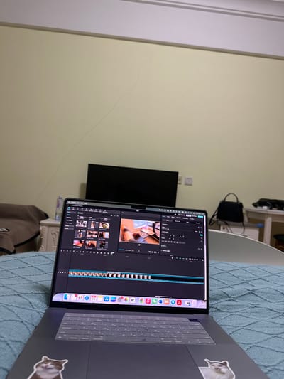 This image depicts an indoor scene in Huangdao District, China, specifically a room, likely a bedroom. In the immediate foreground, a dark gray laptop with its screen open is positioned on a quilted blue bedspread or blanket. The laptop displays video editing software, showing a timeline with numerous clips and a preview window featuring an indoor scene with another laptop, a coffee cup, and an individual's hands. The laptop's keyboard is covered by a translucent protector, and two cat-themed stickers are affixed to the palm rest area. The application dock at the bottom of the screen displays icons for various macOS applications.
In the mid-ground, a light yellow wall dominates, exhibiting a prominent, irregular crack extending diagonally from the upper left towards the lower right. Below this crack, a black, rectangular flat-screen television is visible, positioned against the wall. To the right of the television, two white electrical outlets are mounted on the wall.
To the left of the laptop, a light-colored bedside table or dresser is partially visible, holding a folded brown blanket or throw and a clear plastic bottle. To the right of the laptop, a white table or desk is partially visible, on which a black handbag with a top handle rests, alongside other indistinct dark objects. Part of a white, curved furniture piece, possibly a headboard, is also visible in the lower right background. No individuals are present in the scene. The lighting appears to be artificial, suggesting an indoor environment.