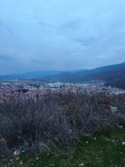 The image depicts an elevated panoramic view of a city, identified as Sveti Konstantin, Bulgaria, at twilight or early evening. The immediate foreground shows a patch of green grass and exposed soil, interspersed with dry vegetation and littered with white plastic bags and other discarded debris. Directly beyond, a dense expanse of bare, dark-branched deciduous shrubs and small trees occupies the lower to mid-frame, with additional white plastic litter caught within its gnarled branches.
The midground reveals a sprawling urban area with numerous buildings, many featuring red-tiled roofs, and scattered artificial lights indicating dusk. Distinct structures include a tall, slender, light-colored minaret-like tower and a prominent industrial chimney rising above the city's rooftops in the central-left area. To the right of the cityscape, a light-colored building with a domed roof is visible on a slight elevation.
The background features multiple layers of mountains, appearing in successively lighter shades of blue-grey as they recede into the distance. The sky above is uniformly overcast with a soft blue-grey hue, indicating a heavily clouded atmosphere or post-sunset conditions. No human figures or discernible actions are present.