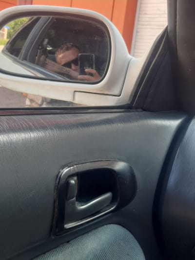 The image is an interior shot of a car's door panel, focusing on the door handle and a side mirror. In the side mirror, a person wearing sunglasses is visible, holding a smartphone and taking a selfie. The person has tattoos on their arm. The car's interior appears to be made of dark plastic and fabric. The exterior background visible through the car window suggests an urban or suburban setting with a building and greenery. The specific location in the city of Shelekhov, Russia is not discernible from the visual information provided.