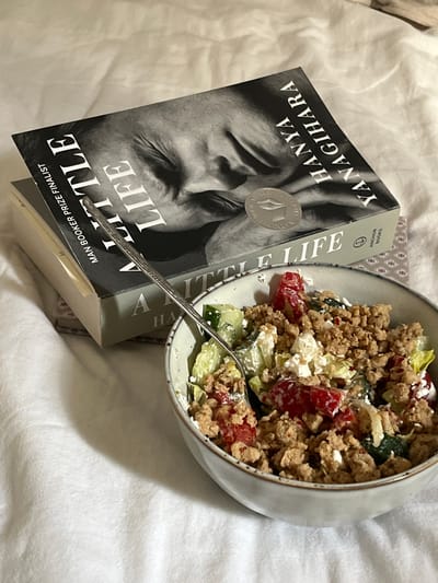 An overhead shot depicts a casual indoor scene on a textured white surface, likely a blanket or duvet. Dominating the upper left is a paperback copy of "A Little Life" by Hanya Yanagihara, identifiable by its title, author, "MAN BOOKER PRIZE FINALIST" text, and a black-and-white cover featuring a person's closed eyes and a hand over their face. A silver circular sticker, possibly indicating an award, is affixed to the cover. Beneath this book, a second, partially obscured book with a patterned cover is visible. To the right, a light grey ceramic bowl with a darker rim contains a mixed dish. The dish consists of crumbled light brown ground meat, diced red vegetables (likely tomatoes), diced green vegetables (likely cucumbers), crumbled white cheese (resembling feta), and some green leafy components (likely lettuce). A silver spoon with an embossed handle rests within the bowl. No people are present. The exact location in the city of Aarhus, Denmark, cannot be determined from the visual information provided.