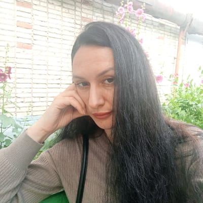 A woman with long, dark hair and light skin poses for a selfie in front of a brick wall. She is wearing a brown, ribbed sweater and has her hand resting on her cheek. Pink hollyhock flowers are visible in the background, along with greenery and a metal pipe. The scene appears to be outdoors, possibly in a courtyard or garden area in the city of Petukhovo, Russia.