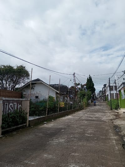 A street scene under a cloudy sky. Dilapidated houses line the road, one with overgrown vegetation. Power lines crisscross overhead. The asphalt street shows signs of wear and tear. A motorcyclist is seen in the distance, driving up the road. The scene appears quiet and somewhat rundown.