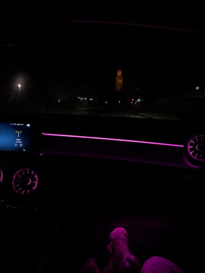 The image shows the interior of a car at night. Purple neon lights illuminate the dashboard and speakers. Outside the window, blurred city lights can be seen. The car's infotainment system is partially visible, displaying radio information. On the right side of the frame, a pair of shoes resting on the passenger side are illuminated in purple.