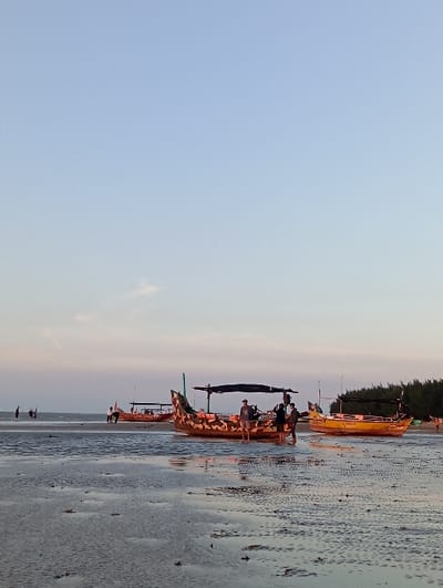 The image depicts a coastal scene at low tide within the city of Tritunggal, Indonesia, under a clear, light blue sky with minimal cloud cover, suggesting either early morning or late afternoon. The foreground consists of wet, textured sand with shallow water pools, reflecting the sky.
Three traditional Indonesian outrigger boats (perahu) are grounded on the exposed sand, with additional boats visible in the distance. The most prominent boat, situated centrally, features a distinct brown and light-colored camouflage-like pattern on its wooden hull and is equipped with a dark canopy. To its right, a bright yellow boat with a similar canopy displays the name "LADIA" painted in black on its hull. A third, darker boat is partially visible to the left and behind the central vessel.
Approximately three to four individuals are gathered near the central boat. One person, dressed in a light-colored top and shorts, stands on the boat's deck. Two other individuals are standing in the shallow water adjacent to the boat, appearing to interact with it or each other. Multiple other human figures are visible in the far distance, walking across the tidal flats or near other distant boats. A line of dark coastal vegetation or trees is visible on the distant right shoreline, marking the edge of the land.