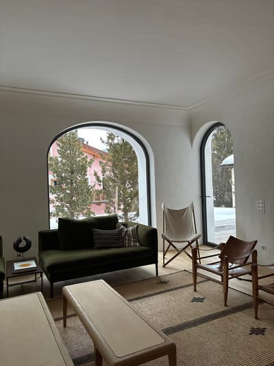 The image shows the interior of a living room in a house located in Celerina/Schlarigna, Switzerland at latitude 46.514, longitude 9.859. The room features a large, green couch placed in front of a large arched window with a view of a snow-covered landscape. The couch is adorned with a throw pillow with a patterned design. To the right of the couch, a wooden armchair with a leather seat and a white canvas chair, folded and placed on the floor, are visible. A brown and white striped rug covers most of the floor in the room. There are two small tables in front of the couch, one of them covered with a book and a black figure. The room has a white ceiling and white walls with a light brown trim. The room is well lit, with natural light streaming in from the arched window. The overall atmosphere is serene and calming.