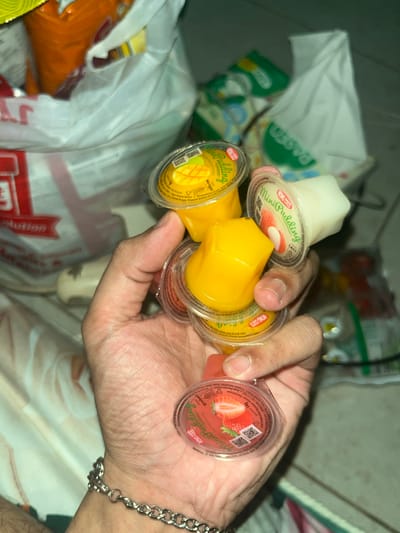 In Meteseh, Indonesia, a close-up shot captures a person's hand holding several small, clear plastic cups of pudding. Three of the cups are distinctly visible. One cup, filled with bright yellow mango-flavored pudding, is held prominently. Another cup, containing white vanilla-flavored pudding, is positioned slightly behind and to the right. A third cup, with red strawberry-flavored pudding, is at the bottom of the frame. The lids of the cups feature branding that reads "Wong Coco Mini Pudding." The hand is adorned with a silver chain bracelet. In the background, partially obscured by the hand and the puddings, are plastic bags filled with various food items, including orange-colored snacks and other packaged goods. The overall setting appears to be indoors, with a soft, diffused lighting.
