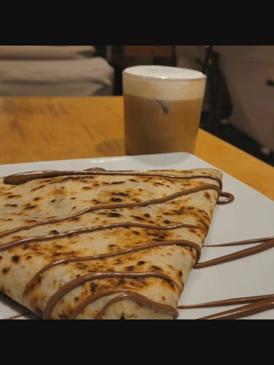 The image shows a close-up view of a folded crepe drizzled with chocolate sauce, presented on a white plate. To the right, a glass of iced coffee with a foamy top is visible. The scene is set on a wooden table, with a blurred background suggesting an indoor cafe or restaurant environment, possibly in Bielawska Wieś, Poland. No people are visible in the frame. The lighting is dim, creating a warm and inviting atmosphere. The focus is on the food and drink, highlighting their textures and presentation.