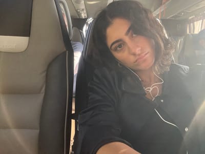 A female individual with dark, wavy, shoulder-length hair and an olive complexion is seated in a bus or coach, looking directly at the camera. She wears a dark collared shirt or jacket with white piping and white wired earbuds, one visible in her left ear. A multi-strand necklace is visible. To her left, the back of an empty passenger seat, upholstered in gray and dark gray/black, displays the faint "Irizar" brand name. The vehicle's interior features overhead lighting fixtures and visible passengers in the background. Bright natural light enters from the right side of the frame, causing a lens flare. The image provides no discernible features to specify a precise location within the city of Pombal, Portugal; the setting is a generic bus interior.