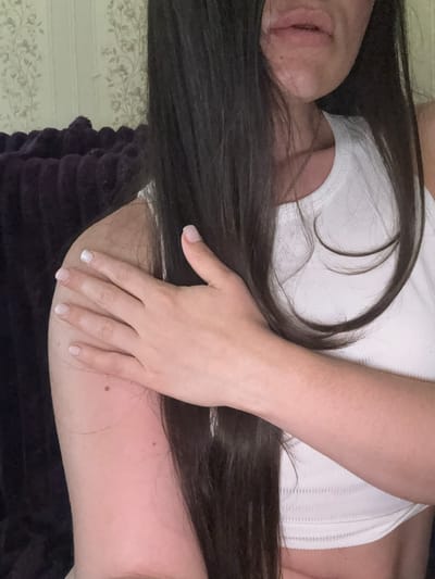A close-up, rotated shot shows a person lying down, their face partially visible with their mouth open. They have long, dark hair that is spread across their chest and arm. The person is wearing a white ribbed tank top. Their right arm is bent, with their hand resting on their upper arm. Their nails are manicured with a light color. The background shows a textured wallpaper with a floral pattern and a dark, plush fabric. The lighting suggests an indoor setting, possibly a bedroom. The overall mood is relaxed and intimate. No text is visible in the image. The location context is Bucha, Ukraine.