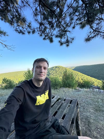 A young man is taking a selfie while sitting on a wooden bench outdoors. He is wearing a black long-sleeved shirt with a yellow graphic design on the front. The man is looking directly at the camera with a neutral expression.
The setting appears to be a scenic overlook in a rural area. In the background, there is a vast expanse of green trees and hills stretching into the distance. The sky is clear and blue, suggesting it is a sunny day. Pine trees with dark green needles are visible in the foreground and frame the left side of the image. The wooden bench he is sitting on is made of weathered planks and has a rustic appearance. There is also some dry grass and rocks surrounding the bench.
The overall mood of the image is relaxed and peaceful, as the man appears to be enjoying the natural scenery. The photo was taken during the daytime, likely in the late afternoon given the soft lighting. There is some visible text on the man's shirt, but it is partially obscured by the graphic and difficult to read clearly. The location is identified as Pryvorottia Druhe, Ukraine.