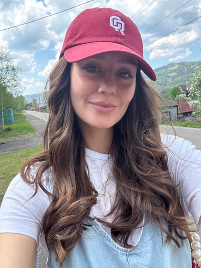 A woman with long, brown hair is posing for a selfie outdoors. She is wearing a burgundy baseball cap with a white logo, a white t-shirt, and a light-wash denim top. The background includes a paved road, grass, trees, and a distant mountain range under a cloudy sky. Based on the background's topography and structures, the location could potentially be Овсянка, Russia.