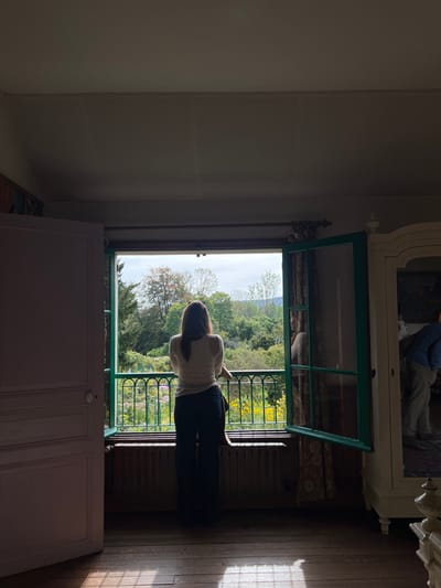 An interior view, likely a bedroom within Claude Monet's house in Giverny, France, shows a female subject standing with her back to the camera, facing an open double window. She has long brown hair and is wearing a light-colored long-sleeved top and dark trousers. Her forearms rest on a dark green ornamental metal railing situated immediately outside the window. The window frames are painted green, and a dark, horizontal radiator is positioned below the window. To the left, a light pink painted wooden door stands partially open. To the right, a tall, ornate white armoire features a mirrored door, reflecting a partial image of another individual, likely male, wearing a light blue top and light-colored pants, in a bent-over posture. The room's interior is dimly lit, with distinct rectangular patterns of bright sunlight cast upon the wooden floor. The exterior view through the window encompasses a vibrant garden filled with numerous yellow and some purple flowers, dense green foliage, and distant trees and hills under a partly cloudy sky.