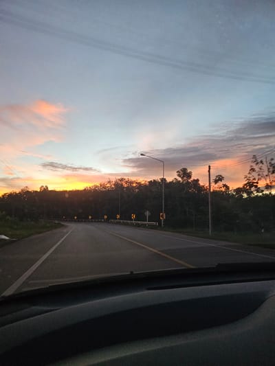 The image captures a scenic view of a road in Khlong Thom, Thailand, during a sunset. The sky is painted with hues of orange, pink, and purple, indicating the sun is setting or rising, casting a warm glow over the scene. Tall trees line the right side of the road, creating a natural barrier and adding to the serene atmosphere. The road itself appears to be a two-lane highway with clear white lane markings, suggesting it's a well-maintained route. Yellow caution signs with black arrows are strategically placed along the roadside, warning of curves ahead. A street light is visible, illuminating the path, which might be useful as dusk settles. The left side of the image is dominated by the sky, with soft, wispy clouds adding texture and depth. A part of a vehicle is visible in the right foreground, suggesting the photo was taken from inside a car, perhaps while traveling along this road. The overall mood is tranquil and picturesque, evoking a sense of peace and the beauty of nature. The time of day is clearly sunset or sunrise, and the location is identified as Khlong Thom, Thailand, through context. There are no people visible in the frame.