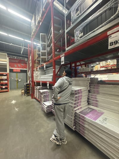 The image shows an aisle in a hardware store, presumably "Prix dépôt" based on the signage. Stacks of packaged doors are visible on the upper shelves, labelled "COOKE LEWIS." A person wearing grey sweatsuit and a black cap, possibly examining the merchandise, is partially in frame. The setting appears to be a warehouse-style retail environment with high ceilings and red metal shelving. Based on the signage "Prix dépôt", it can be assumed the location is in Leers, France.