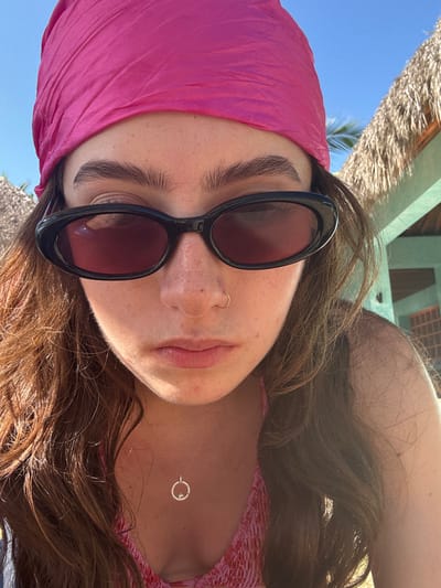 A woman wearing a pink headscarf and black sunglasses is featured in a close-up shot. She has a nose ring and is wearing a delicate necklace with a circular pendant. Her long, brown hair is visible around her face and neck. The background includes a blue sky and a thatched roof structure, suggesting an outdoor setting. The image has a warm, sunny feel.