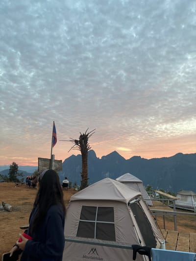 The image depicts a high-altitude camping or viewpoint area in Nong Ian, Laos, at either sunrise or sunset, evidenced by the ambient light and sky colors. The sky is dominated by a dense formation of altocumulus clouds, displaying shades of grey, white, and pale orange-pink at the horizon.
In the far background, a prominent range of dark, jagged mountains with visible valleys is silhouetted against the lighter horizon.
In the midground, from left to right:
* A group of approximately four to five individuals is seated around a low, dark table on a dirt surface, appearing to be engaged in conversation or dining.
* Behind this group, a weathered wooden post with a rectangular sign and the flag of Laos (red, blue, white stripes with a white circle) is visible.
* A tall, slender, dead palm tree with stripped fronds stands centrally.
* An individual wearing a light-colored top is seated alone on a chair, facing the mountain range.
* Multiple beige and white glamping-style tents are visible on raised wooden platforms, connected by wooden walkways.
In the foreground:
* On the left, the back of a person with long, dark hair and a patterned headband is visible. This individual wears a dark long-sleeved top and holds a red object in their right hand and a dark rectangular object (possibly a phone) in their left. Their head is turned slightly to the right, facing towards the tents and mountains.
* On the right, a "Mountainhiker" brand beige tent is prominently positioned on a raised wooden platform. The tent features a rectangular window with a four-pane grid. A black garment and a light blue towel-like item are draped over a railing immediately in front of the tent.
The immediate ground surface is reddish-brown dirt.
