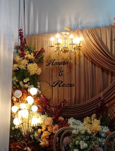 This media file captures a wedding decoration setup in Cibrek, Indonesia. The scene is adorned with an abundance of floral arrangements, featuring a mix of yellow, white, orange, and deep red flowers, along with lush green foliage. A prominent feature is a modern floor lamp with multiple spherical lights on thin stems, illuminating the floral display.

In the background, elegant draping of beige fabric creates a backdrop, accented by a vintage-style chandelier with crystal embellishments hanging from above. The words "Amara & Reza" are elegantly scripted in black font on the fabric, indicating this is a personalized decoration for a wedding ceremony or reception.

The lighting suggests an indoor event, possibly in the evening or during a time when artificial light is preferred. The overall ambiance appears celebratory and romantic, typical of a wedding event. The detailed floral arrangements and personalized text emphasize the festive nature of the occasion.