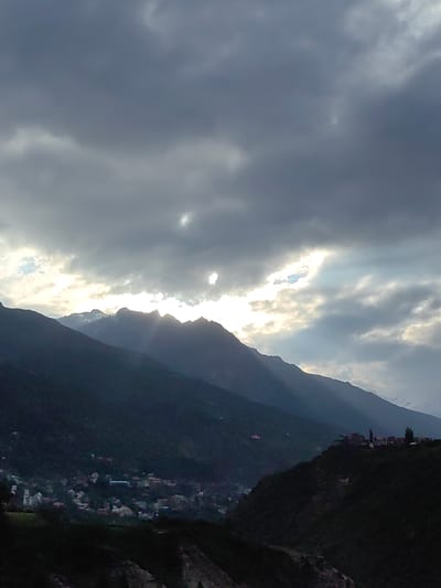 The image depicts a mountainous landscape with a village nestled on a hillside. The sky is overcast with dramatic, dark clouds, but rays of sunlight break through in several places, illuminating parts of the sky and casting shadows on the mountains. The mountains are dark and imposing, with some snow-capped peaks visible in the distance. A village, likely Kardang, India, is visible on the slopes of the mountains. The village appears to be made up of small buildings with red or brown roofs, clustered together. The overall scene is one of natural beauty and tranquility, with a sense of the power and grandeur of nature.