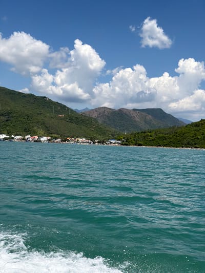 A wide shot captures a coastal scene in Ninh Hòa City, Vietnam. The foreground is dominated by the turquoise waters of the ocean, with white wakes of a moving boat visible at the bottom left. In the midground, a village is nestled along the shoreline, characterized by a collection of buildings with varying roof colors, including red, white, and orange. Fishing boats are moored in the shallow waters near the village. The background features lush, green, mountainous terrain that rises steeply from the coast, with some power lines visible on the slopes. The sky above is a clear blue, dotted with fluffy white clouds.