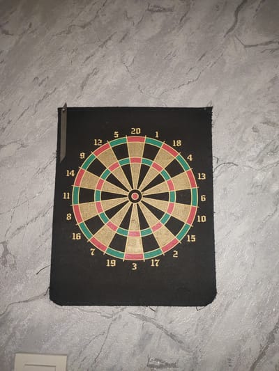 A dartboard is mounted on a textured grey wall. The dartboard is circular with numbered sections. The numbers visible are 20, 5, 12, 9, 14, 11, 8, 16, 7, 19, 3, 17, 2, 15, 10, 6, 13, 4, 18, and 1. The dartboard is black with colored segments in red, green, and gold. There is a small metal object attached to the top left corner of the dartboard. The wall appears to be a roughly textured plaster. The scene is indoors. There are no people or animals present. The image does not contain any information that would allow for determining its location in the city of Esik, Kazakhstan.