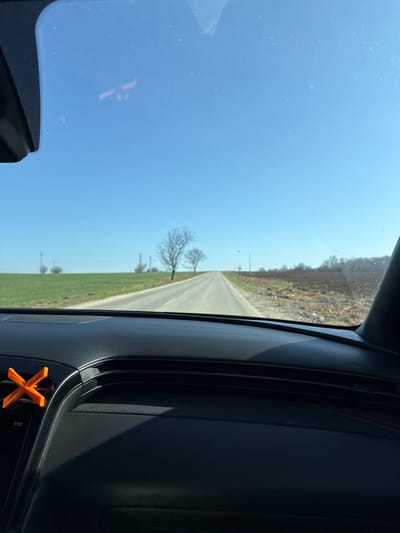 The image depicts a forward-facing view from inside a vehicle traveling on a paved road in Umarevtsi, Bulgaria. The lower third of the frame shows the dark gray dashboard of the vehicle. An orange, X-shaped object, appearing to be a vent clip with the text "TICO," is affixed to an air vent on the left side of the dashboard. Minor reflections and smudges are present on the windshield. The road, appearing to be asphalt and bounded by white solid lines, extends straight into the distance, ascending a slight incline. To the left of the road is a cultivated green field with several bare trees and utility poles. To the right, a reddish-brown, possibly tilled or fallow field is visible, also containing sparse bare trees and utility poles. The sky is clear and bright blue, indicating strong sunlight which casts shadows across the dashboard. No people are visible in the scene.