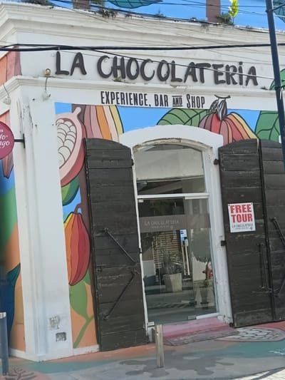 This image shows the exterior of "La Chocolateria" in Puerto Plata, Dominican Republic. The building features a white facade with dark, prominent lettering spelling out "LA CHOCOLATERIA" above a smaller sign that reads "EXPERIENCE, BAR AND SHOP". The storefront has a large glass entrance with dark wooden shutters on either side. A "FREE TOUR" sign is affixed to the right shutter. The building is adorned with a colorful mural depicting cocoa pods and leaves in shades of orange, pink, green, and brown. The ground in front of the building is painted with a colorful, abstract design. The scene appears to be an outdoor commercial establishment.