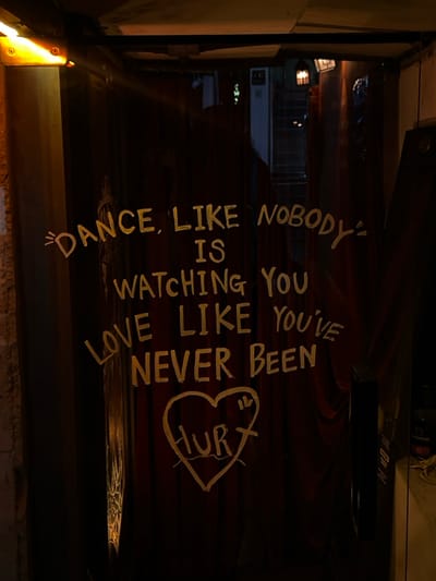 The image depicts a dimly lit, curtained entrance situated in 마포구, South Korea. A dark, possibly maroon, fabric curtain covers the majority of the doorway. Emblazoned on the curtain in light yellow, hand-painted text is the phrase: "DANCE, LIKE NOBODY IS WATCHING YOU LOVE LIKE YOU'VE NEVER BEEN HURT." The word "HURT" is further enclosed within a hand-drawn heart outline. An amber-colored light fixture is affixed to the upper left frame, illuminating a rough-textured wall segment. To the right of the curtain, a dark vertical door handle is visible, and a narrow opening provides a partial view into an interior space. This view reveals what appears to be a white refrigeration unit and several dark-colored bottles on a surface. Above the main curtain, a gap offers a glimpse into a darker internal area, where a lantern-style light fixture and a white wall-mounted appliance, possibly an air conditioner, are visible above a staircase. No individuals are present in the scene.