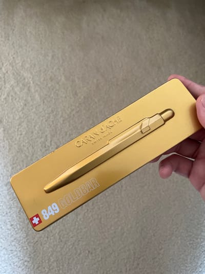 A person's hand holds a golden, rectangular metal tin or presentation box containing a golden ballpoint pen. The box is oriented diagonally, with the top right corner slightly elevated. The pen is embedded in a molded recess within the box, showcasing its hexagonal barrel and distinctive clip. The background appears to be a light-colored, textured surface, possibly carpet or fabric, suggesting an indoor setting.

On the left side of the pen's impression, the brand name "CARAN d'ACHE" is visible, oriented vertically, with "SWISS MADE" printed beneath it. Both texts are debossed or printed in a slightly darker gold tone. On the right side of the pen, also oriented vertically, is the text "849 GOLDBAR" in white lettering. Adjacent to "849," a small red square with a white cross, the Swiss flag, is present. The overall aesthetic is luxurious and highlights the pen as a premium item. The lighting is bright and even, indicative of daytime. The location context is Middle Park, Australia.