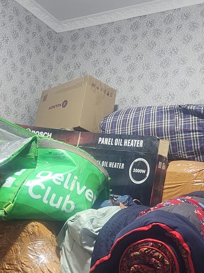 The image depicts a cluttered indoor scene, likely a storage area or a room packed with various items. The dominant colors are muted, with a patterned wallpaper featuring a grey floral design on the wall. In the foreground, a bright green delivery bag with white lettering that reads "Deliver Club" is prominently displayed. Behind the bag, several boxes are stacked. One box has the word "BOSCH" visible on it. Another box is black and clearly labeled "PANEL OIL HEATER" with "2000W" indicated. A large, blue and white plaid blanket or comforter is draped over some of the items. Further in the background, more boxes and rolled-up bedding or textiles are visible. The lighting suggests an indoor, possibly dimly lit environment. There are no people visible in the image. The overall impression is one of accumulated goods. The location is identified as Kara-Köl City, Kyrgyzstan.
