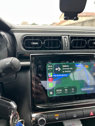 The image depicts the interior of a vehicle, specifically the dashboard and infotainment system, viewed from the driver's or front passenger's perspective. The car's interior appears dusty.

The central infotainment screen displays an active navigation route on a map, indicating the current time as 10:14, with a 5G cellular signal. The navigation prompts the driver to turn left in 200 meters onto "Avenue de la Mairie." Other visible streets on the map include "Avenue de Montbrison" and "Rue de la Becque." The estimated arrival time is 10:30, with 16 minutes remaining for a 17-kilometer journey. This specific configuration of street names places the vehicle in **Saint-Cyprien, Loire, France**.

Above the screen are three horizontal air vents, with the central vent featuring a vertical adjustment scroll. A black plastic phone mount is visible adhered to the windshield or upper dashboard, suggesting a mobile device may have recently been or is about to be secured.

Below the infotainment screen, a row of control buttons and a rotary knob are visible. The front defroster button is illuminated with an orange indicator light, suggesting it is active. A red triangular hazard light button is also present. To the lower left, a set of car keys, attached to a blue lanyard, hangs partially visible. A small white rectangular object, possibly a toll pass transponder, bearing the "VINCI" logo, is positioned near the lower controls.

Through the windshield, an overcast sky is visible, along with the indistinct outline of light-colored buildings in the distance, consistent with the exterior environment of Saint-Cyprien, France. A faint reflection of a hand or arm is visible on the lower left portion of the infotainment screen.
