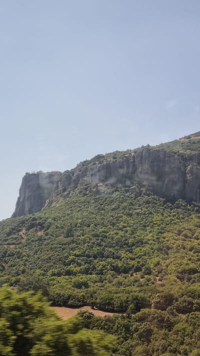 This image depicts a vertical view of a mountainous landscape near the city of Kalambaka, Greece. The upper portion of the image is dominated by a clear, pale blue sky. The middle section features a dramatic, sheer rock face, with a dense, green forest covering its lower slopes and extending into the foreground. Atop the prominent rock formation on the left, several structures, possibly part of a monastery or settlement, are visible. The rocky cliffs exhibit distinct strata and weathered textures. The forested area is lush and varied, with a mix of deciduous and coniferous trees. In the foreground, the greenery appears blurred, suggesting motion, possibly from a vehicle passing by. The overall scene conveys a sense of natural grandeur and historical presence, characteristic of the Meteora region.