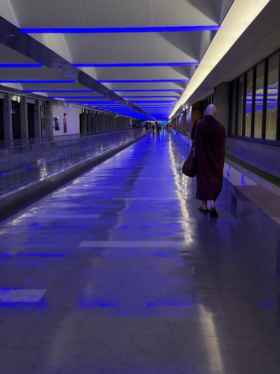 A photograph captures a man in a maroon robe walking along a corridor illuminated by blue lights. The corridor walls are lined with square panel sections divided by vertical blue lighting. The floor surface reflects the blue lights. In the distance, there are figures and lights, indicating the depth of the corridor. Above, a dark ceiling structure is visible. Based on the available context, the photo was taken in Taoyuan City, Taiwan, but no specific location information is provided.