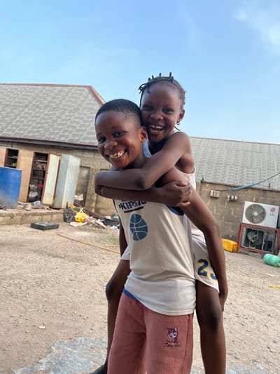 Two children are photographed outside in Iddah, Nigeria. One child carries the other on their back. Both children are smiling broadly. The child on the bottom is wearing a light-colored basketball jersey and maroon pants with a cartoon character on the leg. The child on top is wearing a light-colored tank top. The background shows corrugated metal rooftops, a brick building with an air conditioning unit, and a dirt ground with scattered trash and debris. The sky is visible in the upper left corner.