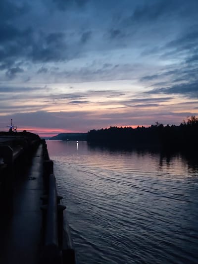 The image depicts a tranquil scene at dusk or dawn along a body of water, likely a river or canal, near the village of Badozhskoi Pogost, Russia. The sky is filled with dramatic clouds, transitioning from deep blues and grays at the top to soft pinks and oranges near the horizon. A dark, silhouetted treeline borders the far bank of the water. On the left side of the frame, a dark, elevated walkway or deck juts out, with a railing and a navigational pole or antenna visible. The water's surface is rippled, reflecting the muted colors of the sky. There are no people visible in the image.