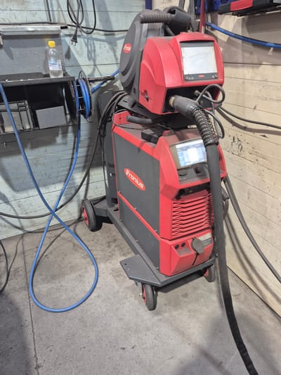 This image depicts a red and gray Fronius welding machine, a piece of industrial equipment, positioned on a concrete floor. The machine is mounted on a wheeled cart and is connected to various cables. In the background, a light-colored wooden wall with horizontal planks is visible, along with some metal shelving and a bottle of water. The scene appears to be an indoor workshop or industrial setting, possibly located in Ylämylly, Finland, given the context. There are no people or specific actions depicted in the image, only the static arrangement of the welding equipment and its surroundings.
