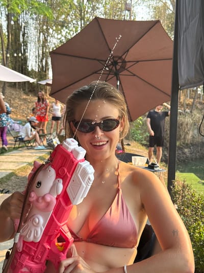 The image depicts a woman holding a pink and white water gun with a unicorn design, firing water droplets towards the camera. She is wearing sunglasses and a pink top. In the background, under a brown umbrella, a group of people are seated. Further back, a man in a black outfit stands. The setting appears to be an outdoor space, possibly in Tambon Nam Phrae, Thailand.