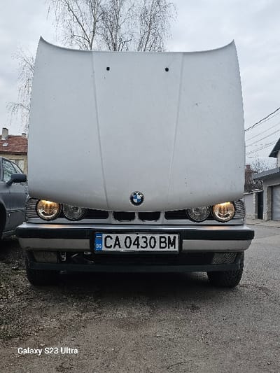 The scene depicts a silver-colored BMW sedan parked on a gravel and asphalt surface in Dobroslavtsi, Bulgaria. The vehicle's hood is fully open and propped vertically. Its front headlights are illuminated, emitting a warm yellow light. A Bulgarian license plate, "CA 0430 BM," is centrally mounted on the front bumper. To the left of the BMW, a portion of a dark-colored vehicle is visible, followed by a building with a reddish-brown tiled roof. To the right, a building with a dark roof and a closed garage door is present, with overhead utility lines visible in the upper right. Bare tree branches are observed against an overcast sky in the background. No individuals are present in the frame. The image was captured with a Galaxy S23 Ultra device.