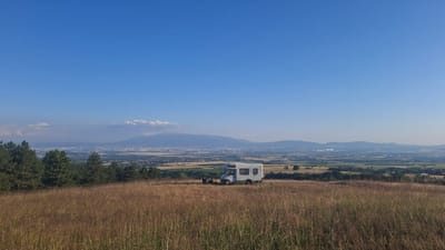 A wide-angle landscape photograph captures a serene vista under a clear blue sky. In the foreground, a field of tall, dry grass stretches across the frame, its golden hues illuminated by the sun. A white camper van is parked on a gentle slope within this field, positioned slightly to the right of the center. Next to the camper van, two dark figures, likely people, are seated, suggesting a moment of rest or observation.
Beyond the grassy field, a line of dark green evergreen trees forms a natural border, their dense foliage contrasting with the dry grass. In the mid-ground, a sprawling city is visible, its buildings appearing as a low-lying urban expanse. This city is situated in a valley, with a distant mountain range forming the backdrop. The mountains are shrouded in a light haze, their contours softened by the distance and atmospheric conditions. The overall impression is one of tranquility and a remote, natural setting overlooking a populated area. The image captures the location in the vicinity of Balsha, Bulgaria.