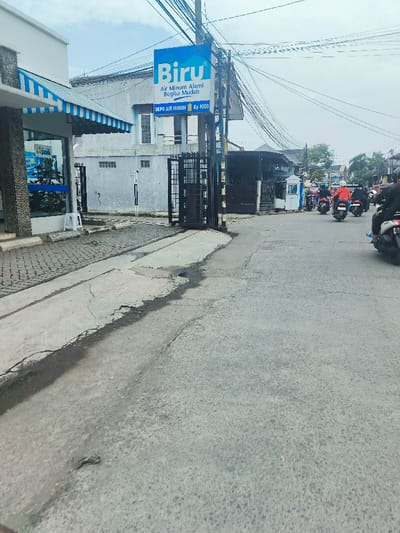 The image shows a street in Kecamatan Dayeuhkolot, Indonesia, located at latitude -6.963 and longitude 107.596. The street is paved with gray asphalt and has a few cracks and patches. There is a large sign for "Biru" water, which reads "Air Minum Alami Begitu Mudah" and "Depo Air Minum Rp 8000." There are a few buildings along the street, including a small shop with a blue awning and a larger, gray building with a black gate. There are four people riding motorcycles in the distance. The sky is overcast and gray. There are numerous power lines running across the sky.