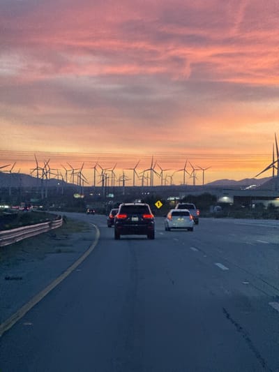 The image captures a highway scene in Desert Hot Springs, United States, under a vivid sunset sky. In the foreground, vehicles are travelling along the road, including a dark SUV in the middle lane and a white car in the right lane. Further in the distance, there are wind turbines dotting the horizon, indicating the presence of a wind farm. A pedestrian crossing sign is visible on the right side of the road. The sky displays a gradient of colors, ranging from orange to pink, as the sun sets. The overall environment suggests a twilight setting on a highway near a renewable energy installation.
